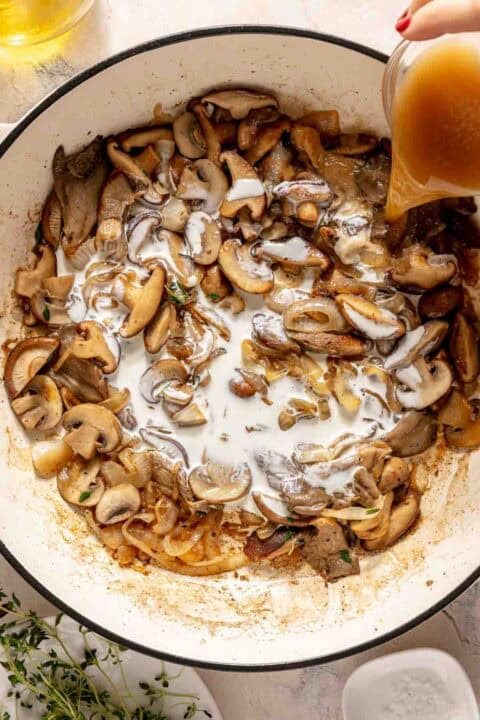Sliced mushrooms and onions sautéing in a skillet as a hand pours broth into the pan.