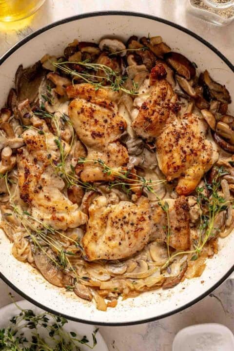 Chicken breasts cooked with mushrooms, onions, herbs, and cream sauce in a skillet, garnished with sprigs of thyme.