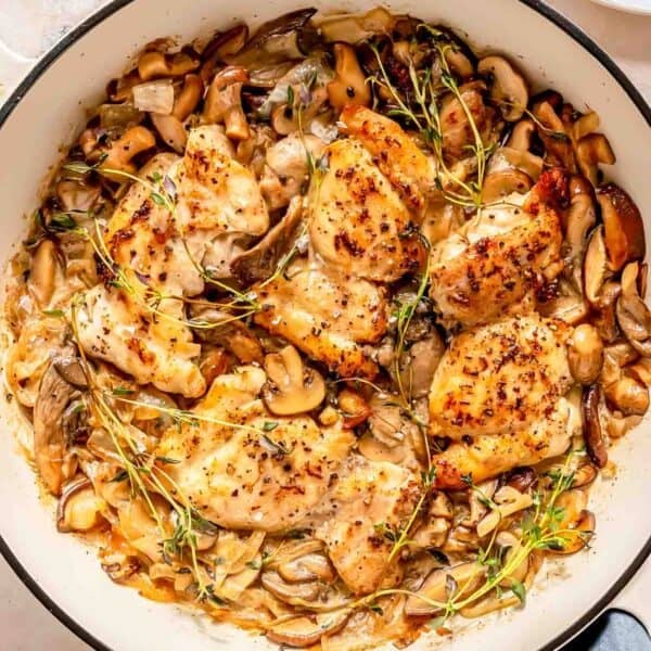A skillet filled with cooked chicken thighs, mushrooms, onions, and fresh herbs, topped with sprigs of thyme.