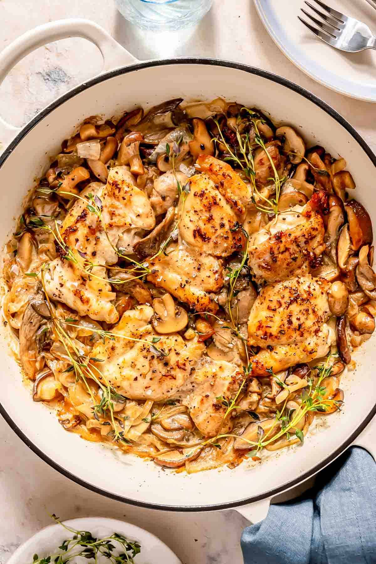 A skillet filled with cooked chicken thighs, mushrooms, onions, and fresh herbs, topped with sprigs of thyme.