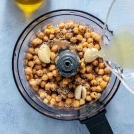 A food processor bowl filled with chickpeas, garlic, spices, and a lemon juice being poured in, with a bottle of olive oil nearby on a light blue surface.