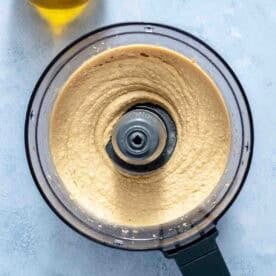 Overhead view of a food processor filled with smooth, blended hummus next to a bottle of olive oil on a light surface.