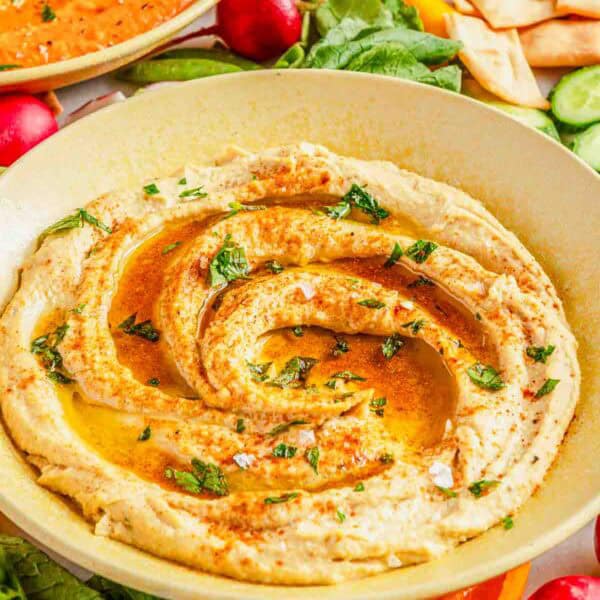 A bowl of garlic hummus topped with olive oil, herbs, and spices, surrounded by fresh vegetables and pita chips.