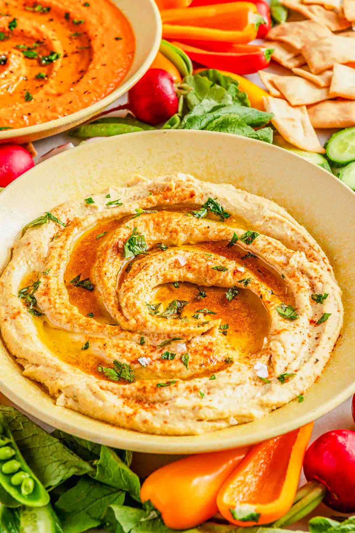A bowl of garlic hummus topped with olive oil, herbs, and spices, surrounded by fresh vegetables and pita chips.