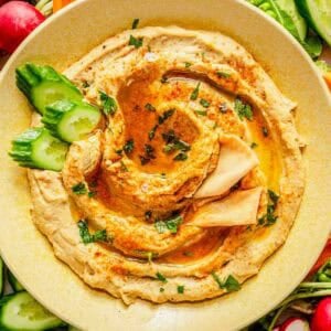 A bowl of garlic hummus garnished with olive oil, paprika, and chopped herbs, served with sliced cucumber, pita, and assorted fresh vegetables.