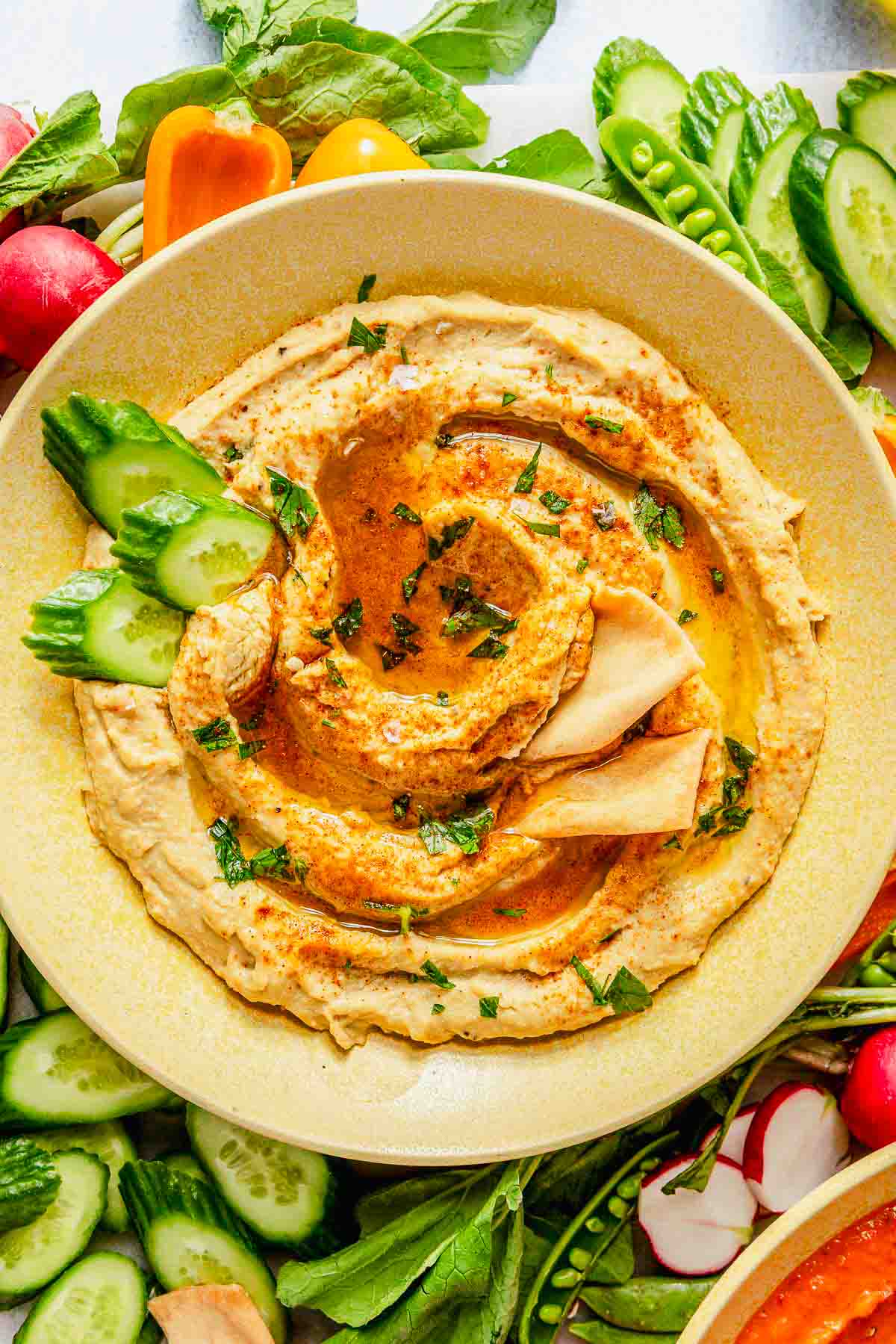 A bowl of garlic hummus garnished with olive oil, paprika, and chopped herbs, served with sliced cucumber, pita, and assorted fresh vegetables.