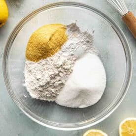 A glass bowl containing flour, granulated sugar, and cornmeal sits on a countertop surrounded by lemons, a sliced lemon, and a metal whisk with a wooden handle.