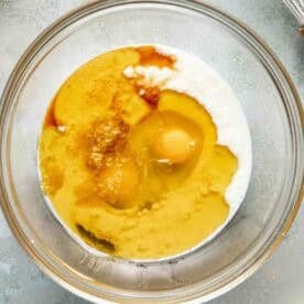 A clear glass bowl containing eggs, milk, vanilla extract, and oil, with a metal whisk and lemon slices nearby on a gray surface.