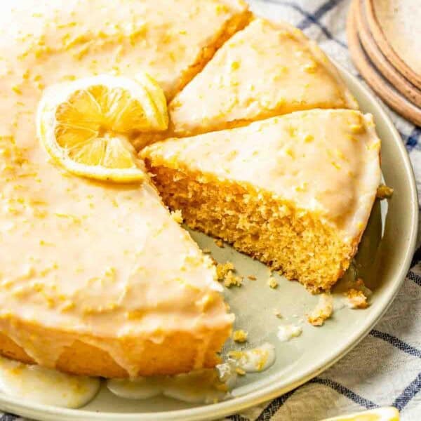 A round lemon cake with a glazed icing and lemon slice garnish, partially sliced on a plate, sits on a checkered cloth with lemons and dishes nearby.