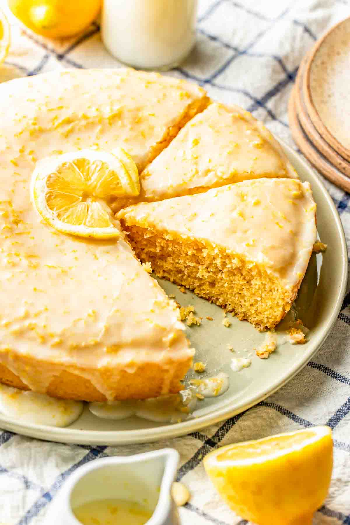 A round lemon cake with a glazed icing and lemon slice garnish, partially sliced on a plate, sits on a checkered cloth with lemons and dishes nearby.