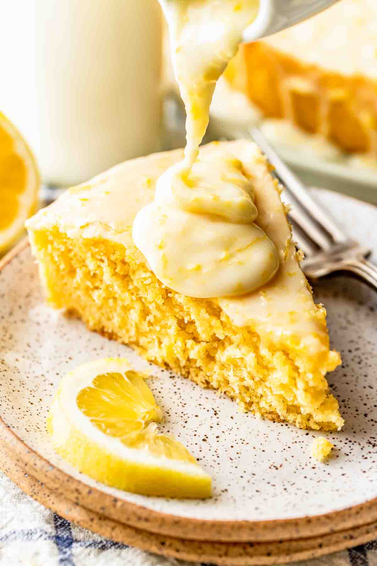 A slice of lemon cake on a plate is topped with thick lemon icing, with a lemon wedge and a fork beside it.