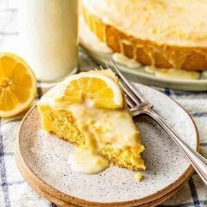 A slice of lemon cake with icing and a lemon slice on top, served on a plate with two forks, next to a bottle of milk and a halved lemon.