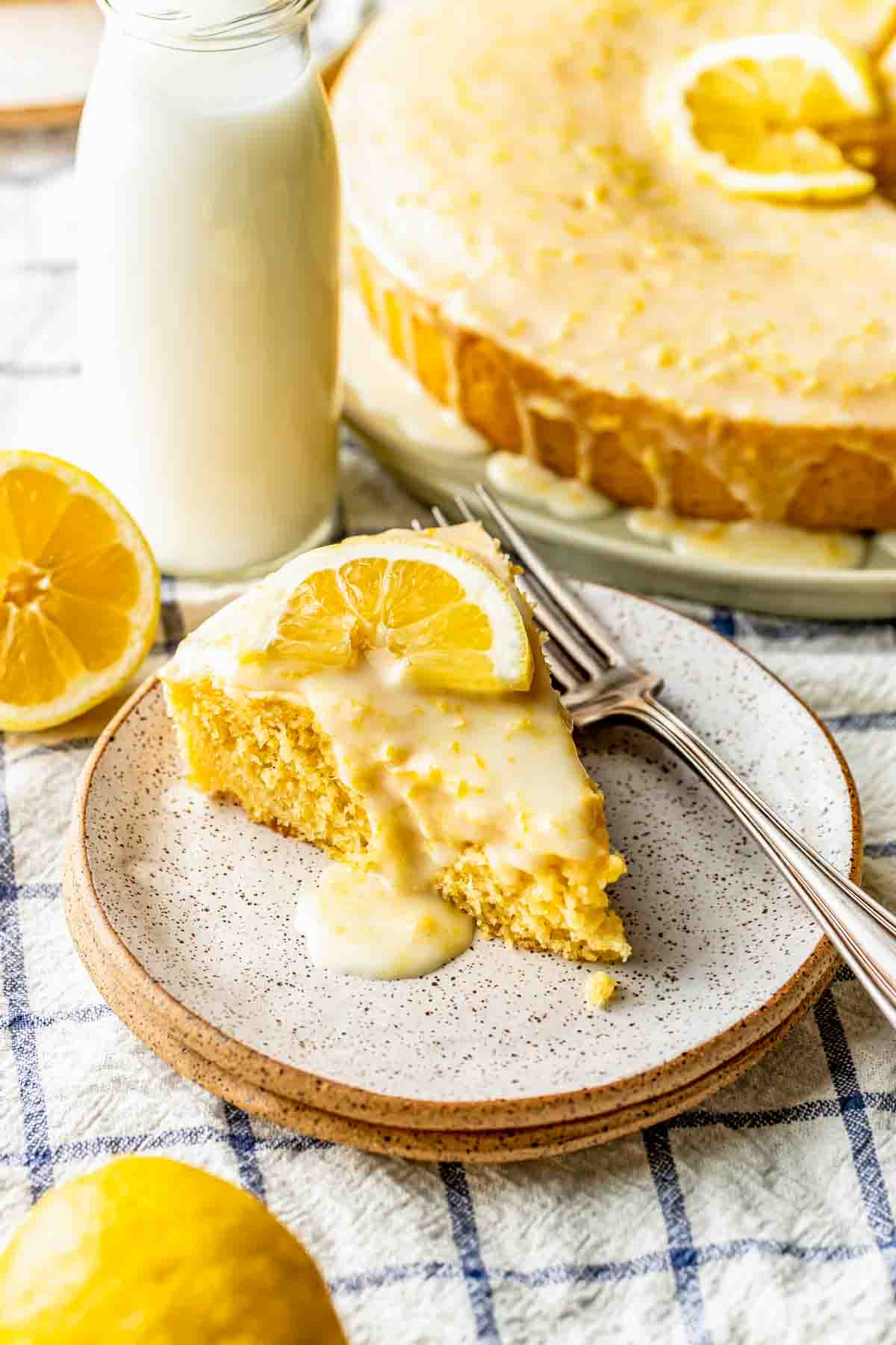 A slice of lemon cake with icing and a lemon slice on top, served on a plate with two forks, next to a bottle of milk and a halved lemon.