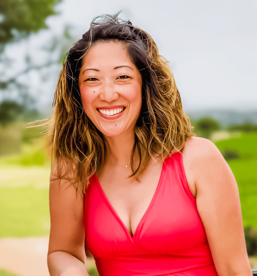 Julie Chiou of Table for Two with wavy brown hair and a bright pink sleeveless top smiles outdoors, with greenery and a tree in the blurred background.