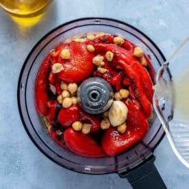 A food processor bowl filled with roasted red peppers, chickpeas, and garlic, with liquid being poured in; a jar of olive oil is nearby.