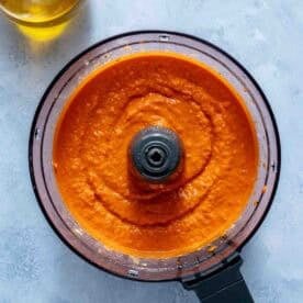 A food processor filled with blended red pepper hummus sits on a light gray surface next to a glass container of olive oil.