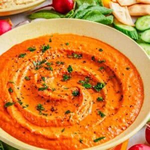 A bowl of red pepper hummus is garnished with herbs and surrounded by fresh vegetables and pita chips.