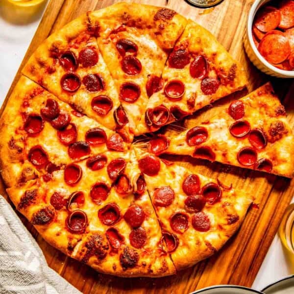 A sliced pepperoni pizza on a wooden board with a pizza cutter, a bowl of pepperoni slices, and drinks nearby.