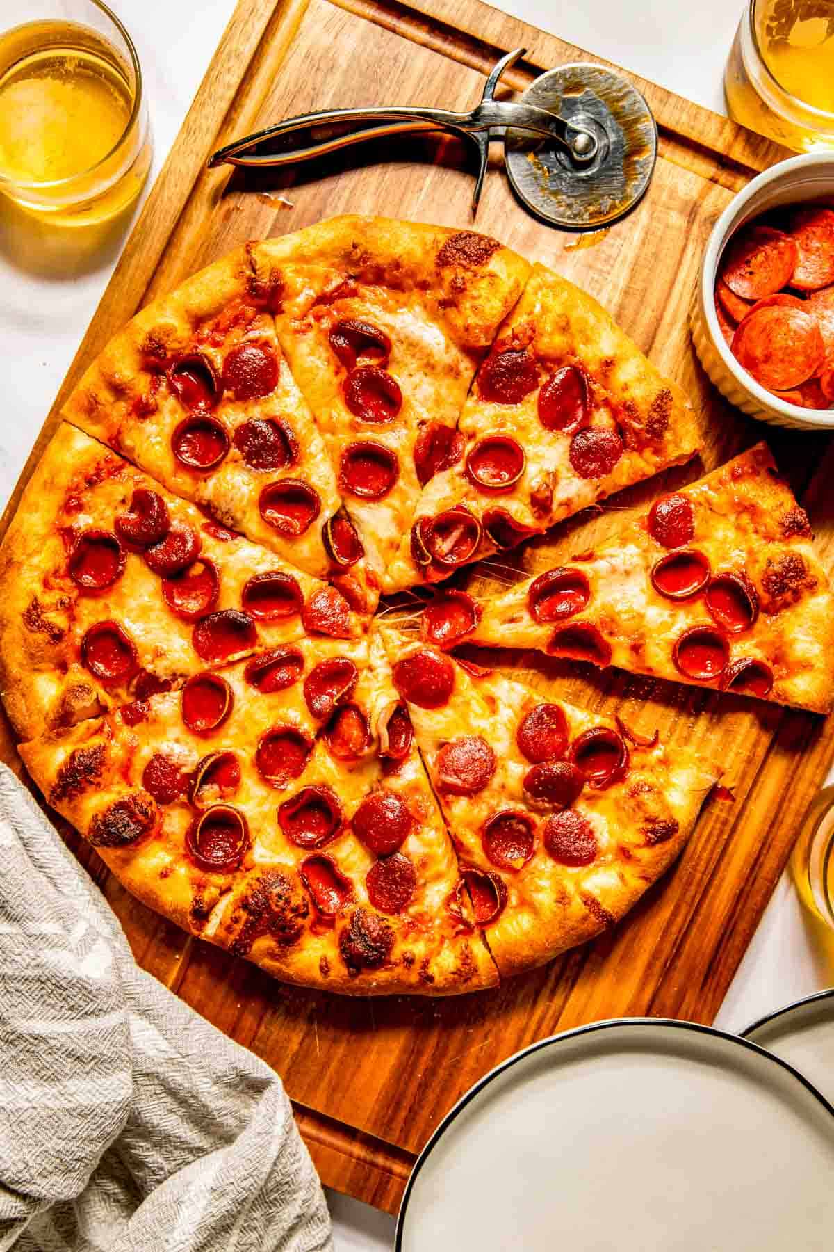 A sliced pepperoni pizza on a wooden board with a pizza cutter, a bowl of pepperoni slices, and drinks nearby.
