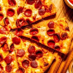 A sliced pepperoni pizza on a wooden cutting board with a bowl of extra pepperoni on the side.