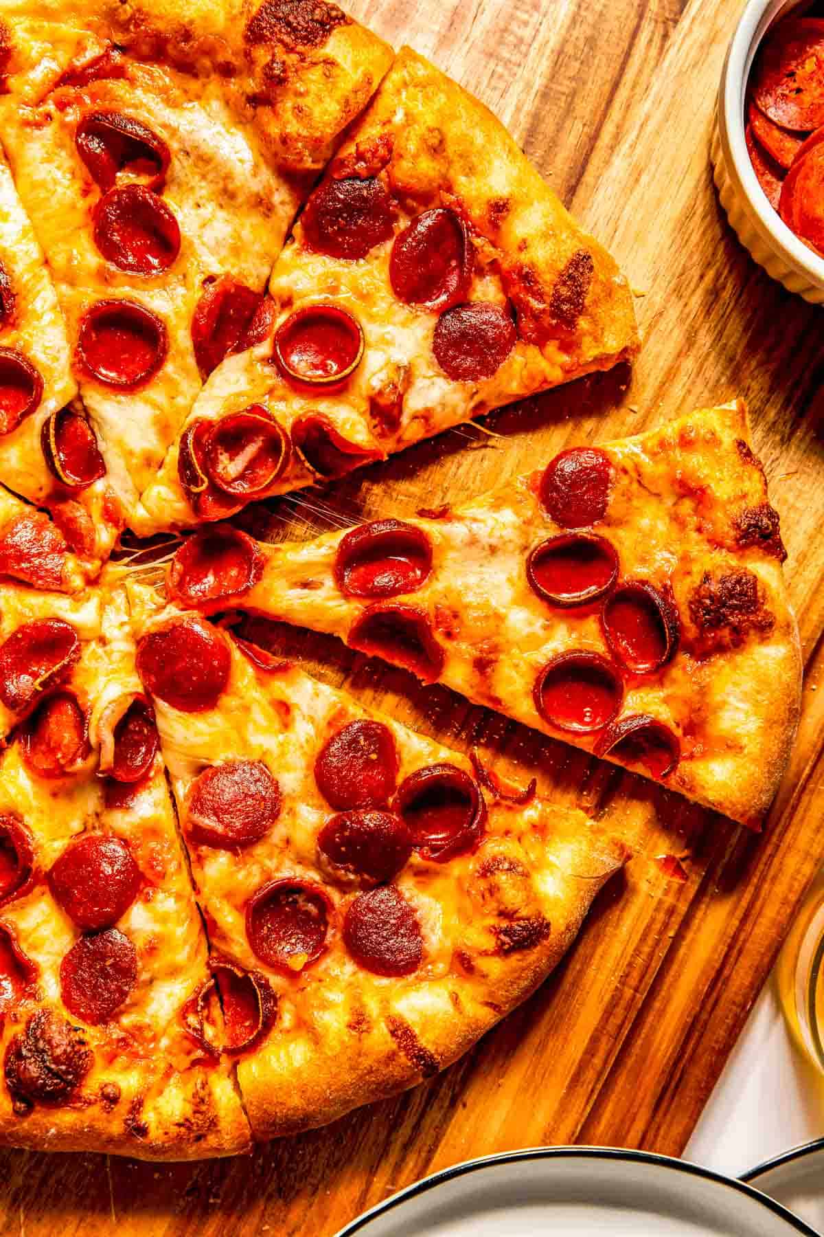 A sliced pepperoni pizza on a wooden cutting board with a bowl of extra pepperoni on the side.