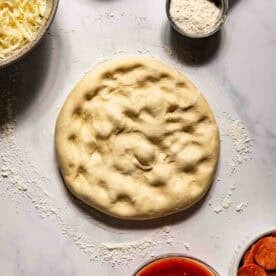 Round pizza dough on a floured surface, surrounded by bowls of shredded cheese, flour, cornmeal, pizza sauce, and sliced pepperoni.