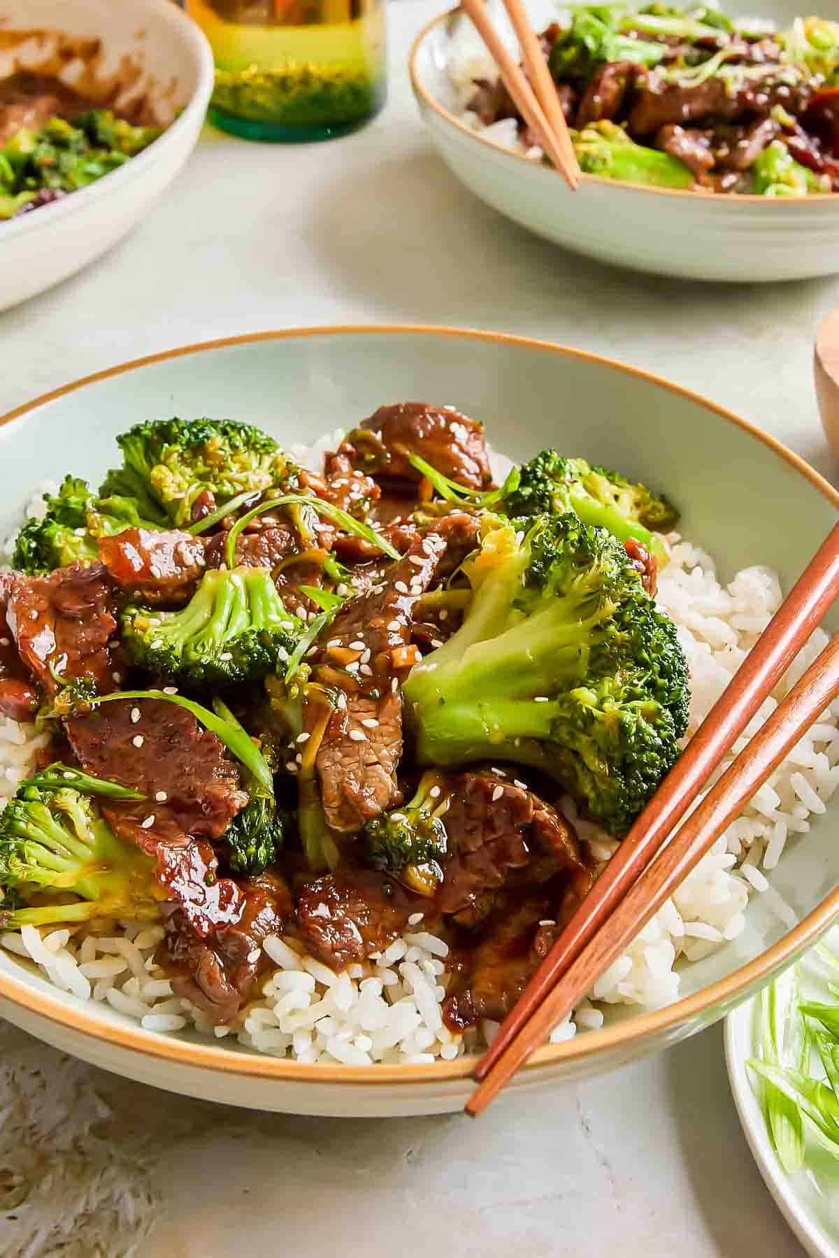 A bowl of white rice topped with beef, broccoli, and teriyaki sauce, garnished with sesame seeds, with wooden chopsticks resting on the bowl.