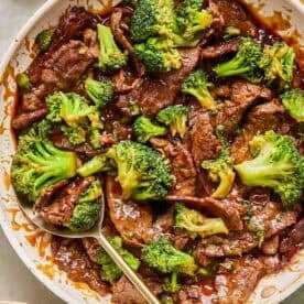 A skillet filled with teriyaki beef and broccoli stir-fry, with a serving spoon and a bowl of white rice nearby.