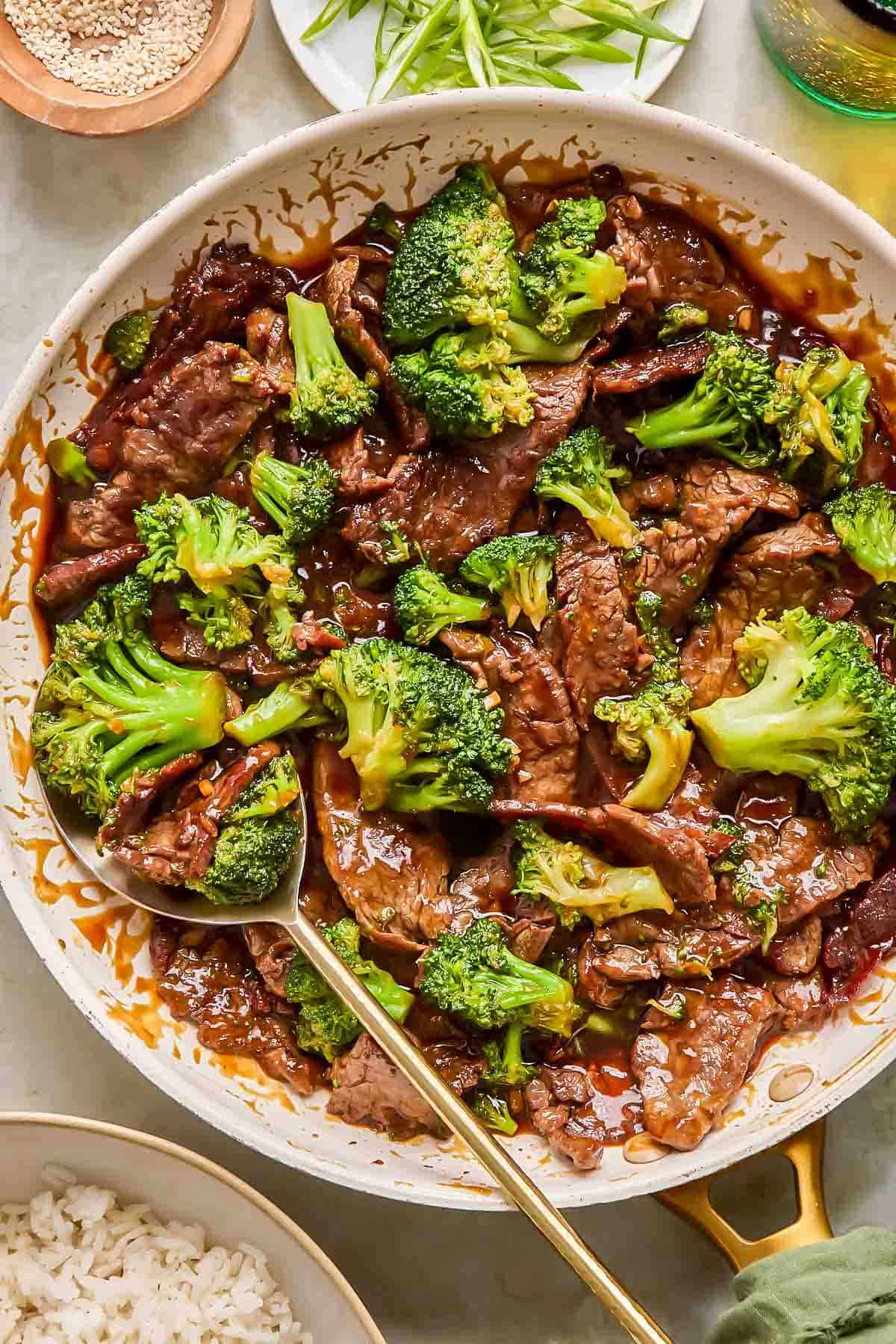 A skillet filled with teriyaki beef and broccoli stir-fry, with a serving spoon and a bowl of white rice nearby.