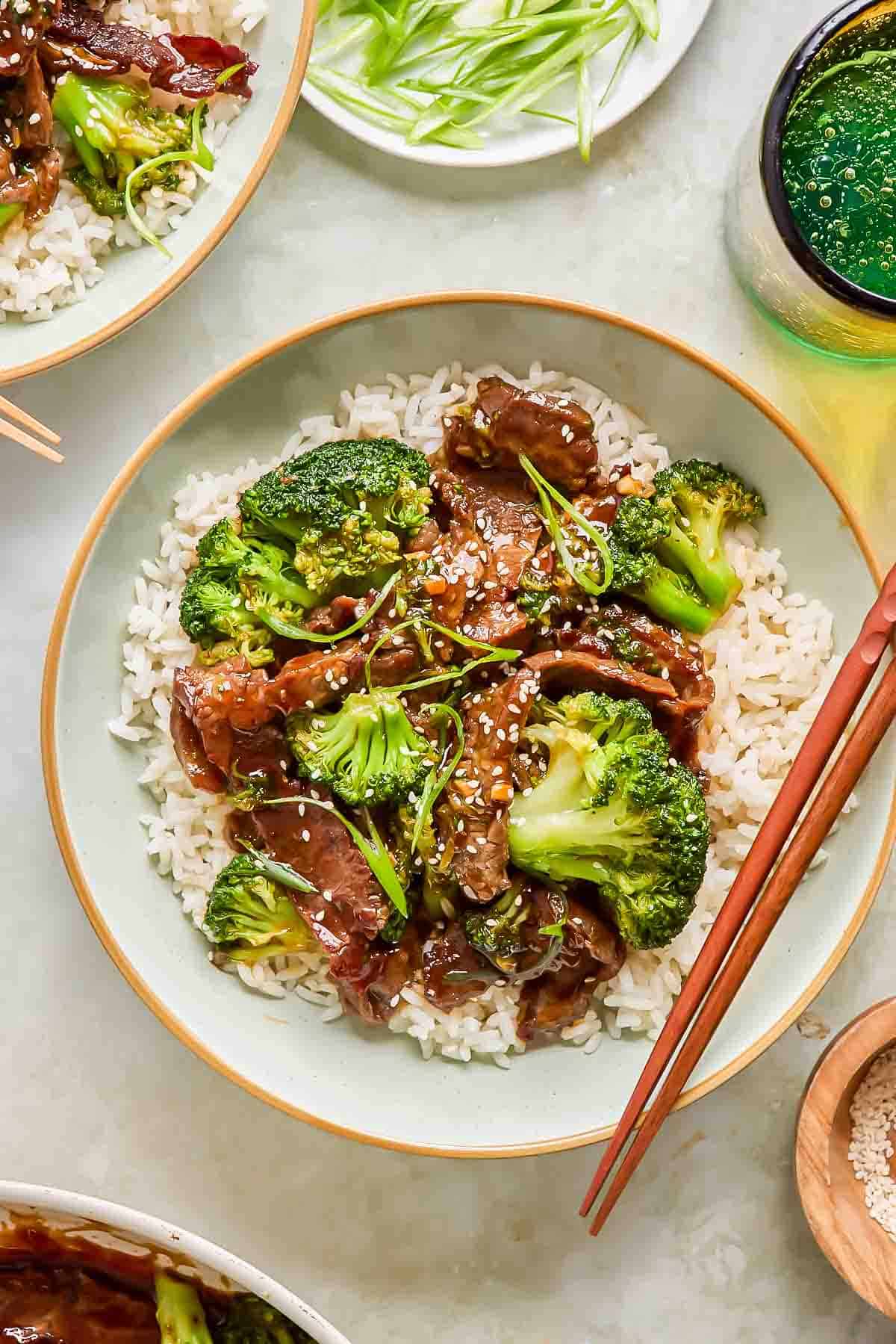 A bowl of white rice topped with broccoli florets and beef in teriyaki sauce, garnished with sesame seeds, with chopsticks resting on the bowl.
