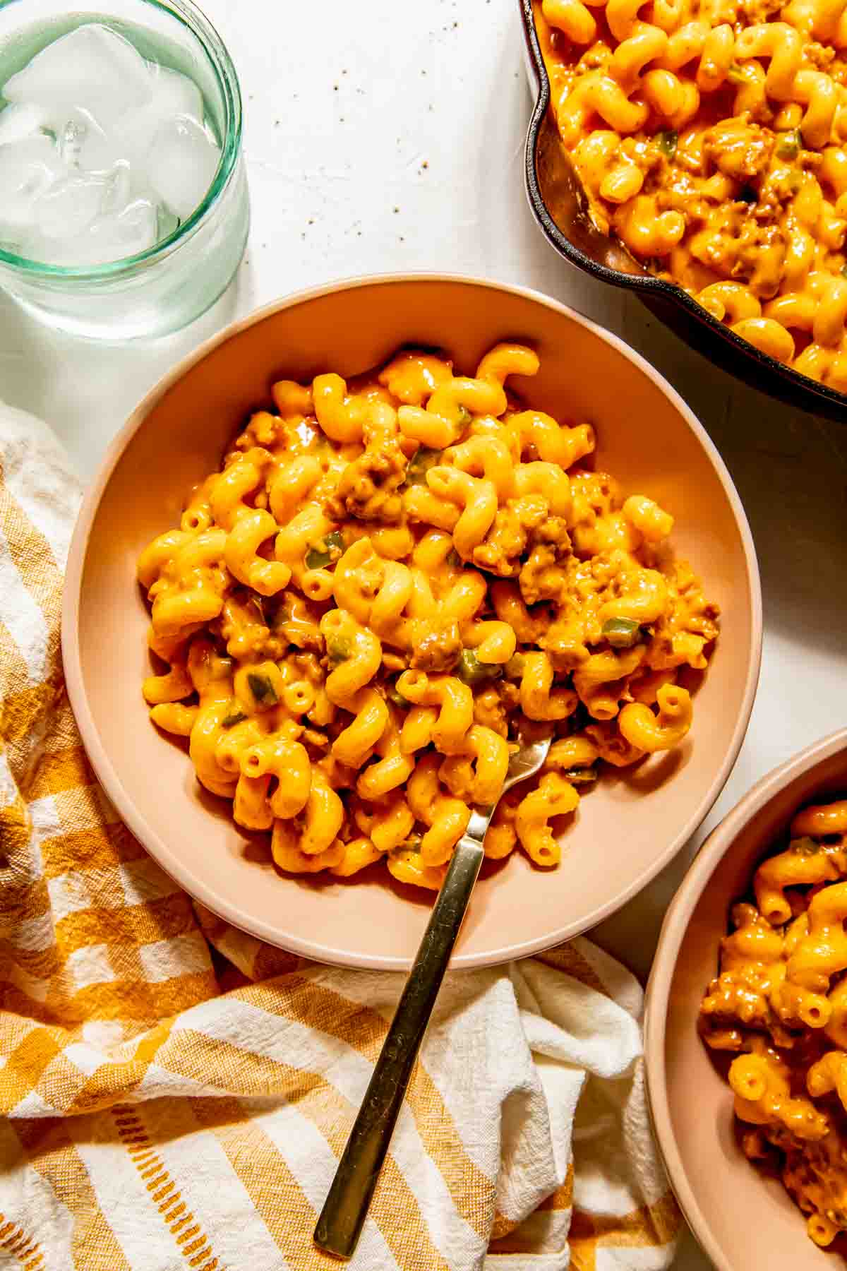 A bowl of chorizo macaroni and cheese sits on a striped towel, with a fork in the bowl and a glass of ice water nearby.