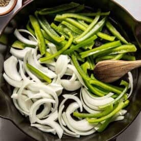 Sliced green bell peppers and onions in a cast iron skillet with a wooden spoon, surrounded by small bowls of salt and pepper.