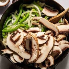 Sliced green bell peppers, onions, and portobello mushrooms in a cast iron skillet with a wooden spoon, surrounded by small bowls of salt and pepper.