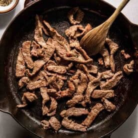 Strips of beef cooking in a cast iron skillet with a wooden spoon, surrounded by small bowls of salt and pepper.