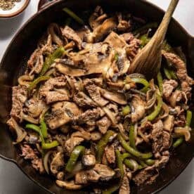 A cast iron skillet filled with cooked beef strips, sliced mushrooms, green bell peppers, and onions, with a wooden spoon resting inside. Bowls of salt and pepper are nearby.