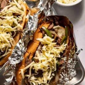 Two cheesesteak sandwiches in foil, topped with shredded cheese, sliced beef, and green peppers, with a bowl of extra shredded cheese nearby.