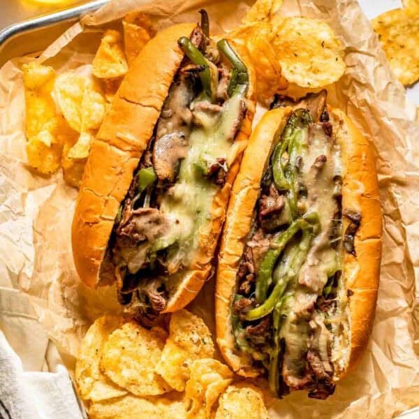 Two cheesesteak sandwiches with melted cheese, sliced beef, and green peppers on hoagie rolls, served with potato chips on parchment paper.