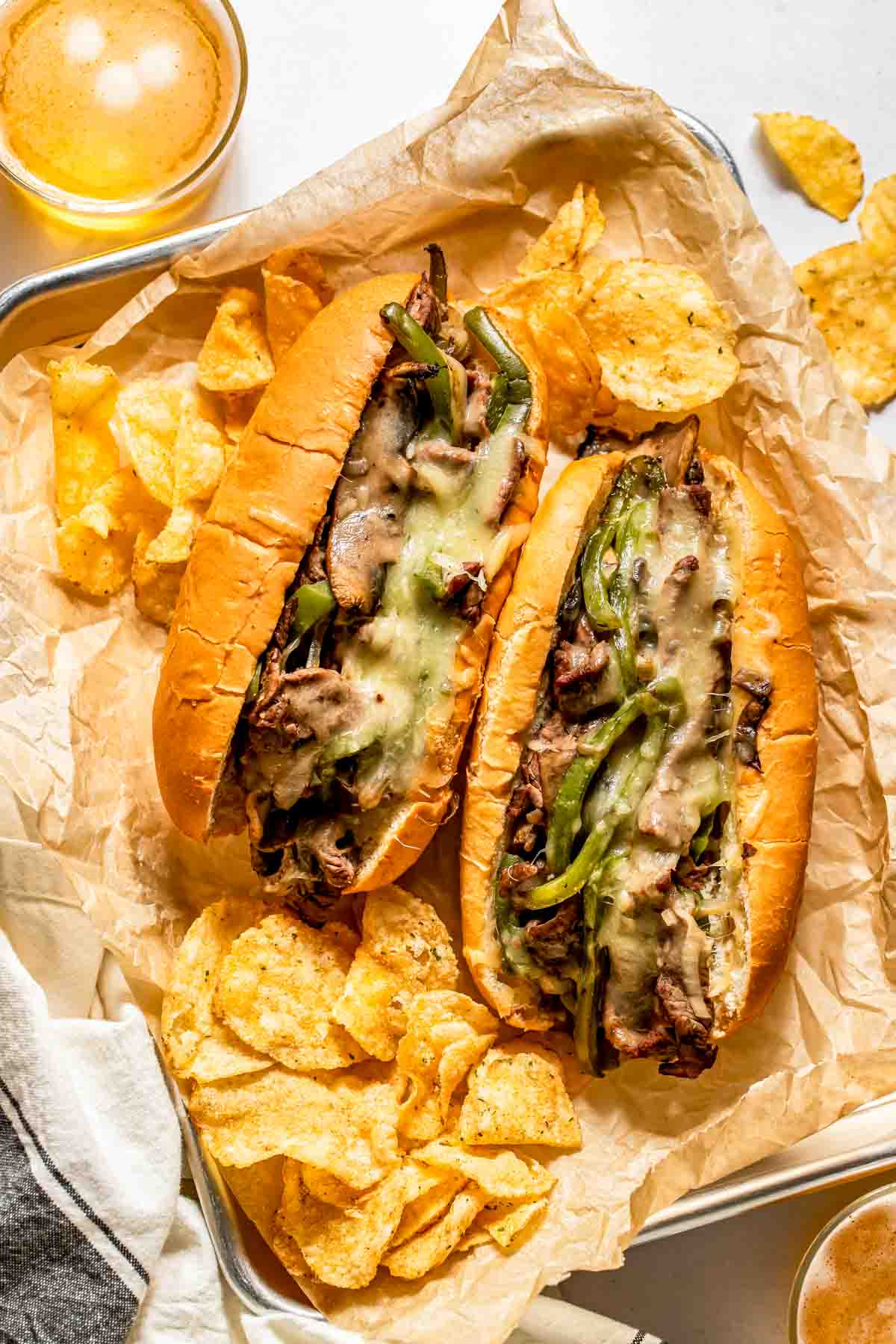 Two cheesesteak sandwiches with melted cheese, sliced beef, and green peppers on hoagie rolls, served with potato chips on parchment paper.