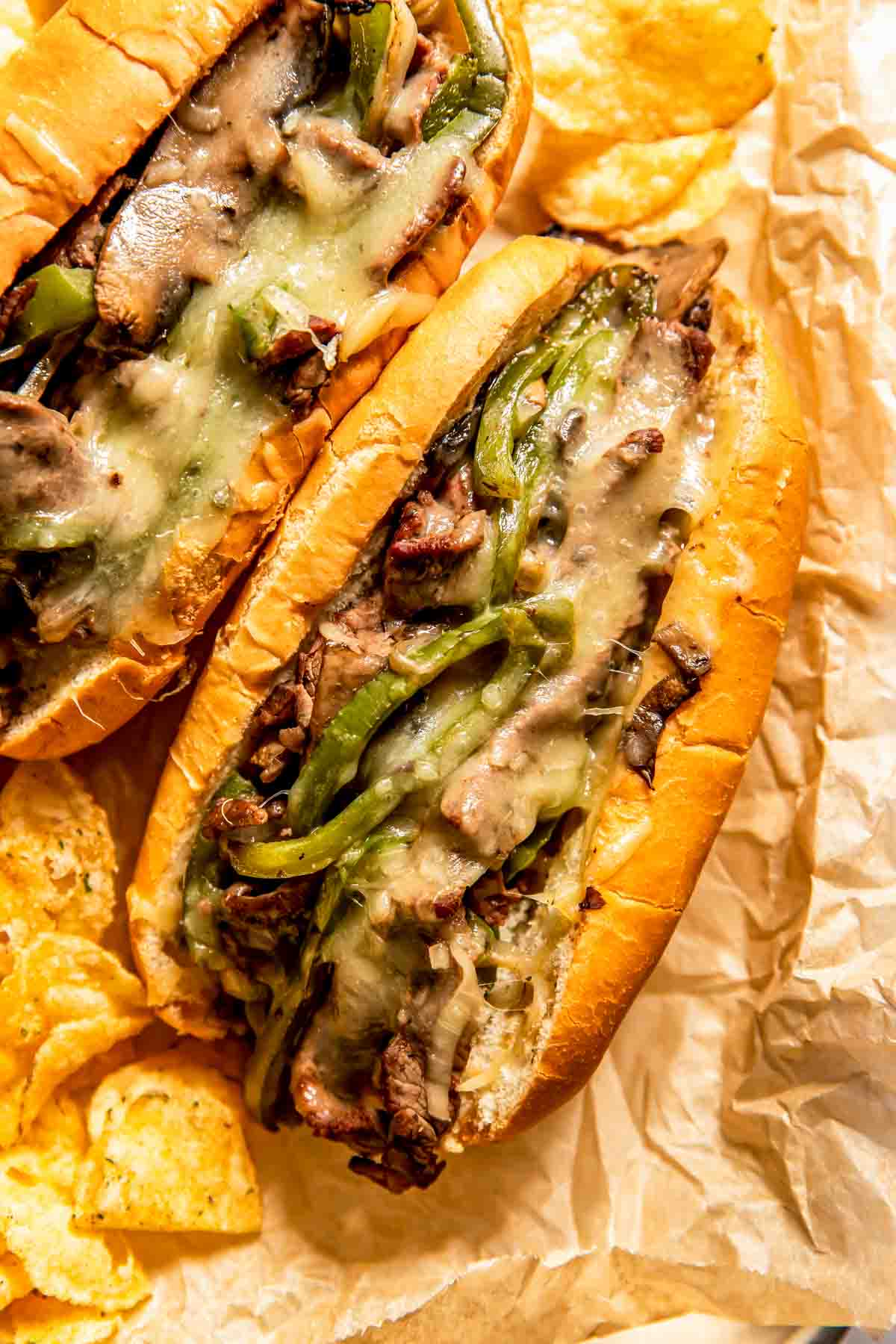 Two Philly cheesesteak sandwiches filled with sliced beef, melted cheese, mushrooms, and green peppers, served on parchment paper with potato chips.