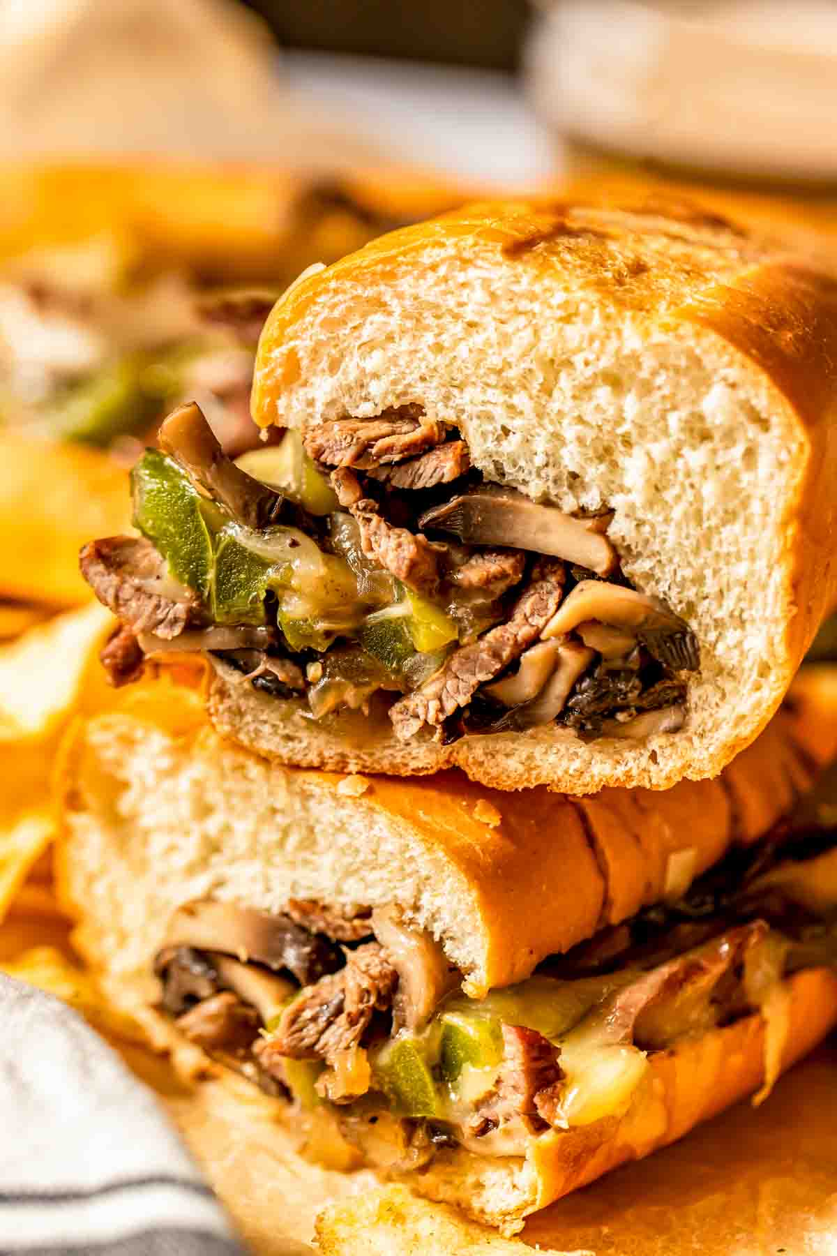 A close-up of a sandwich filled with sliced beef, melted cheese, mushrooms, and green bell peppers, cut in half and stacked.