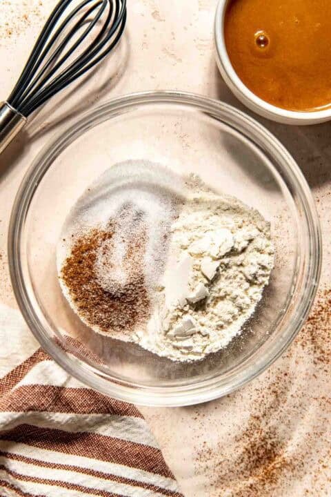 A glass bowl with flour, sugar, cinnamon, and salt on a countertop, next to a whisk, a bowl of salted caramel sauce, and a striped towel.