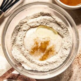 A glass bowl with flour, sugar, oil, vanilla extract, and milk, next to a bowl of salted caramel sauce and a whisk on a light countertop with a striped towel.
