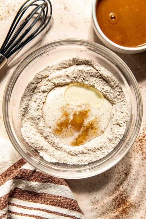 A glass bowl with flour, sugar, oil, vanilla extract, and milk, next to a bowl of salted caramel sauce and a whisk on a light countertop with a striped towel.