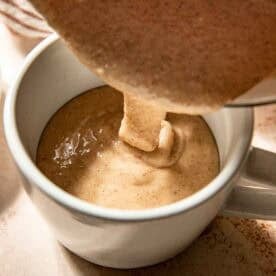 Thick, cinnamon-spiced batter being poured from a bowl into a white mug on a light surface.