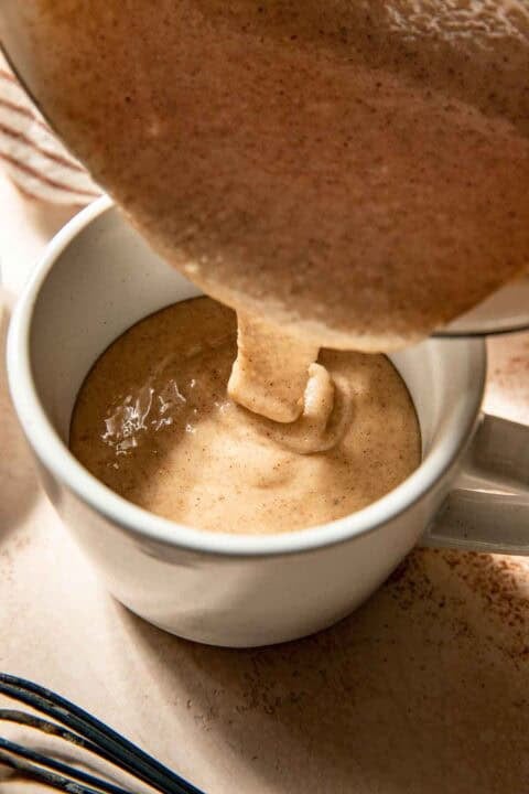 Thick, cinnamon-spiced batter being poured from a bowl into a white mug on a light surface.