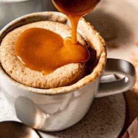 A spoon pours caramel sauce over a mug cake in a white mug placed on a speckled plate with a spoon beside it.