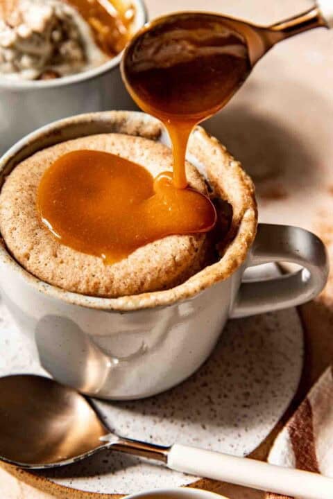 A spoon pours caramel sauce over a mug cake in a white mug placed on a speckled plate with a spoon beside it.