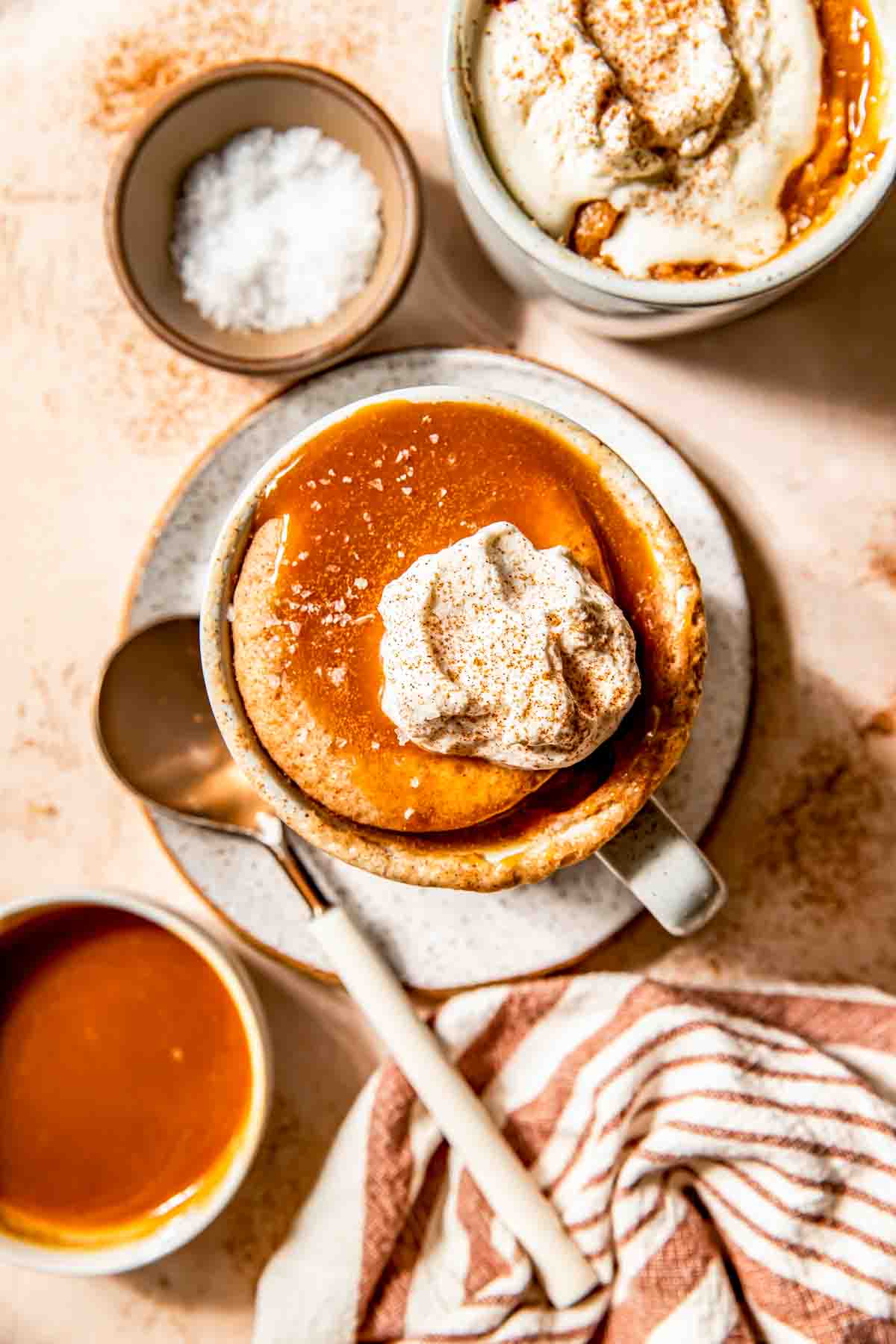A mug of caramel dessert topped with whipped cream and cinnamon sits on a plate with a spoon; a bowl of salted caramel sauce, a bowl of salt, and a striped napkin are nearby.