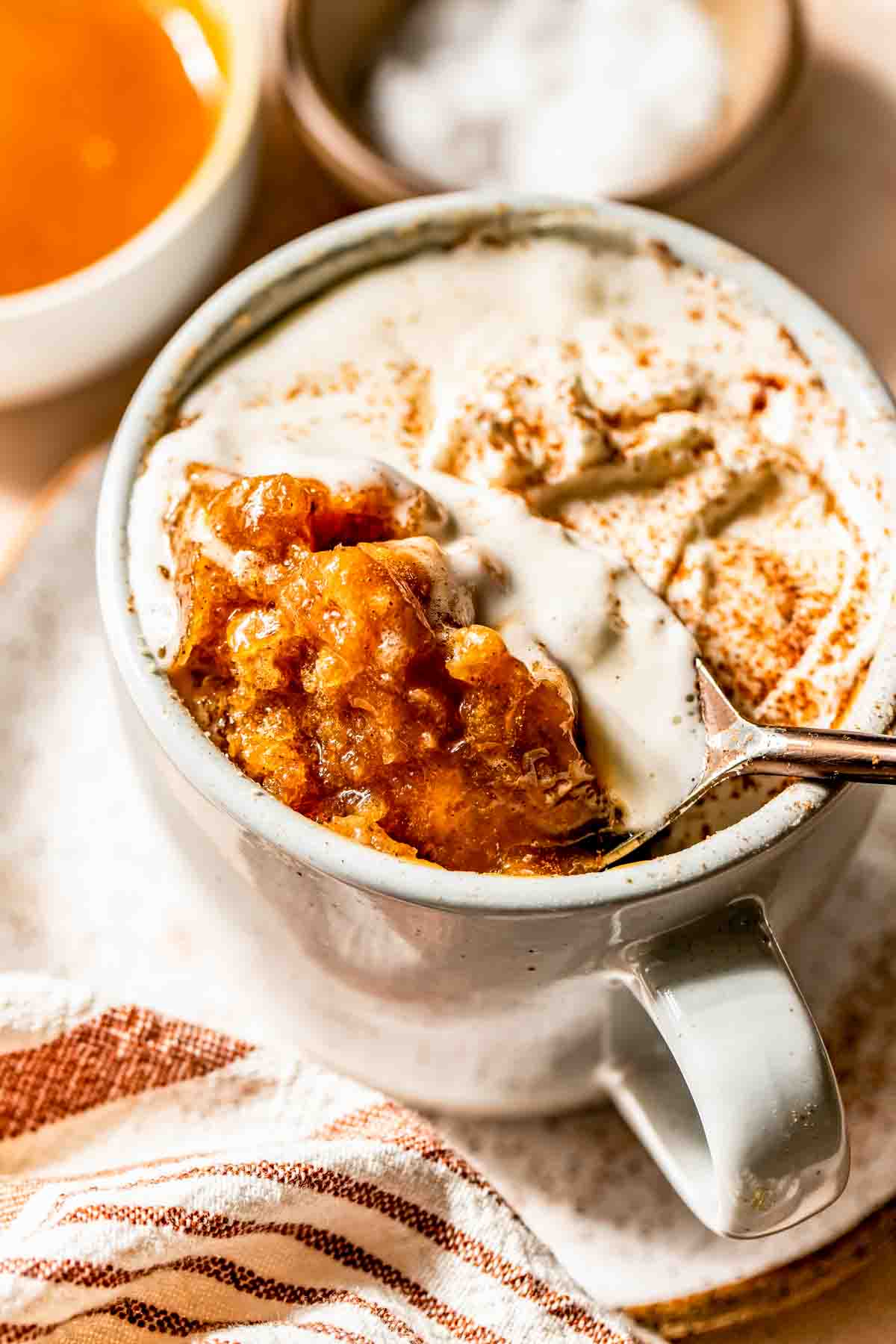 A mug filled with a baked dessert topped with creamy sauce, with a spoon lifting a portion; a striped napkin and bowls are nearby.