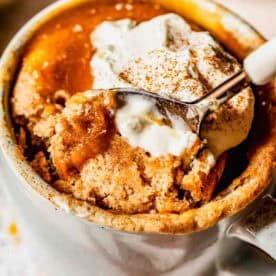A close-up of a mug filled with cinnamon spiced mug cake, topped with a dollop of whipped cream and a sprinkle of cinnamon, with a spoon scooping out a portion.